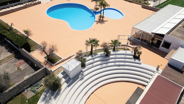 An aerial view of a swimming pool with a curved deck, surrounded by palm trees and a building with a sloped roof. - Amor di Salento by BarbarHouse (San Foca)