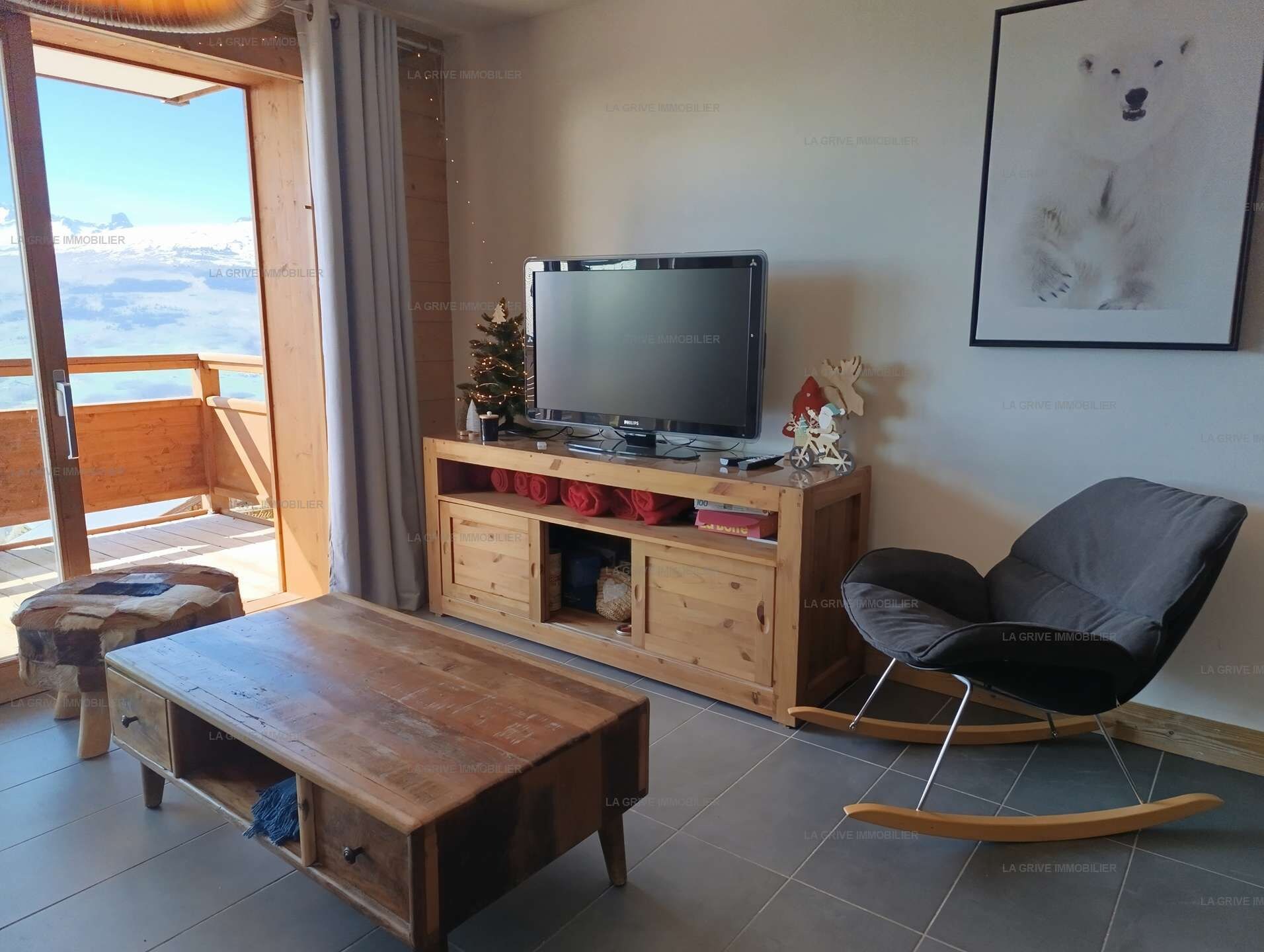 Un salon avec une table basse en bois, un téléviseur posé sur un meuble en bois, un fauteuil à bascule et un tableau encadré accroché au mur.