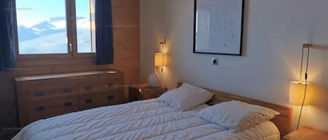 A bedroom with a bed, a dresser, a nightstand, a lamp, a framed picture, and a window with a view of mountains.