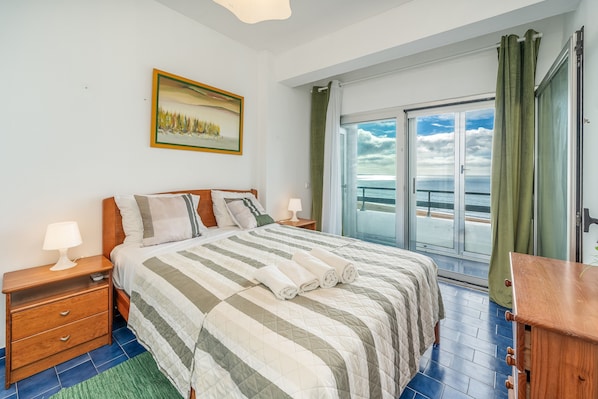 A bedroom with a bed, nightstands, a painting, a view of the sea through a glass door, and a wooden dresser.