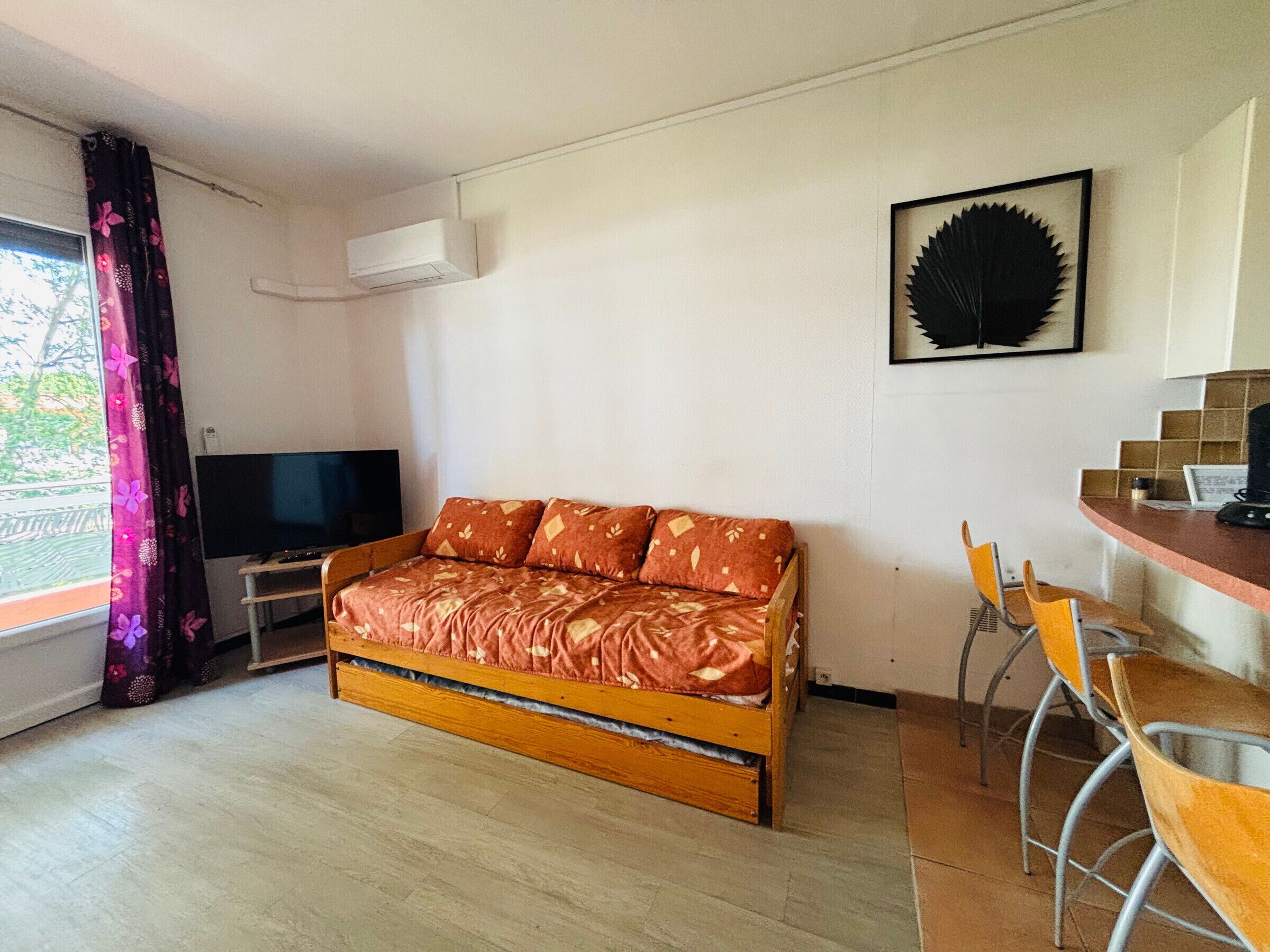 Espaço compacto para viver, com sofá, televisão, área de refeições com cadeiras e janela com cortinas.