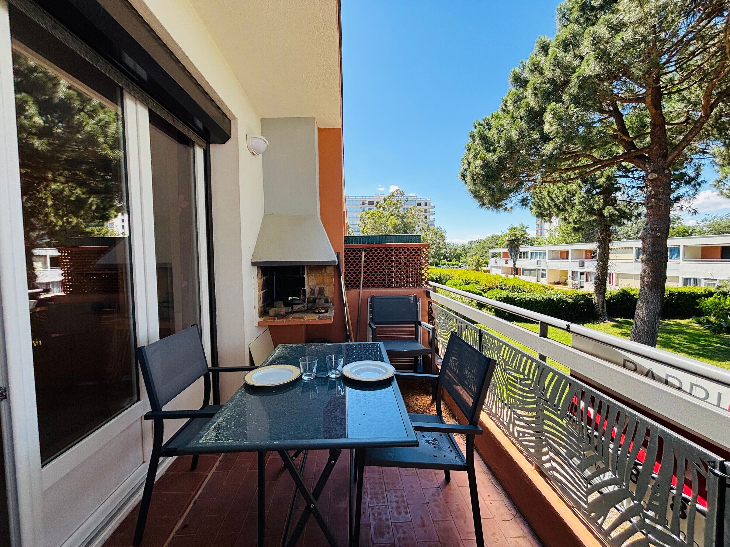 Un balcon avec une table et des chaises, un barbecue et une vue sur les bâtiments et les arbres environnants.