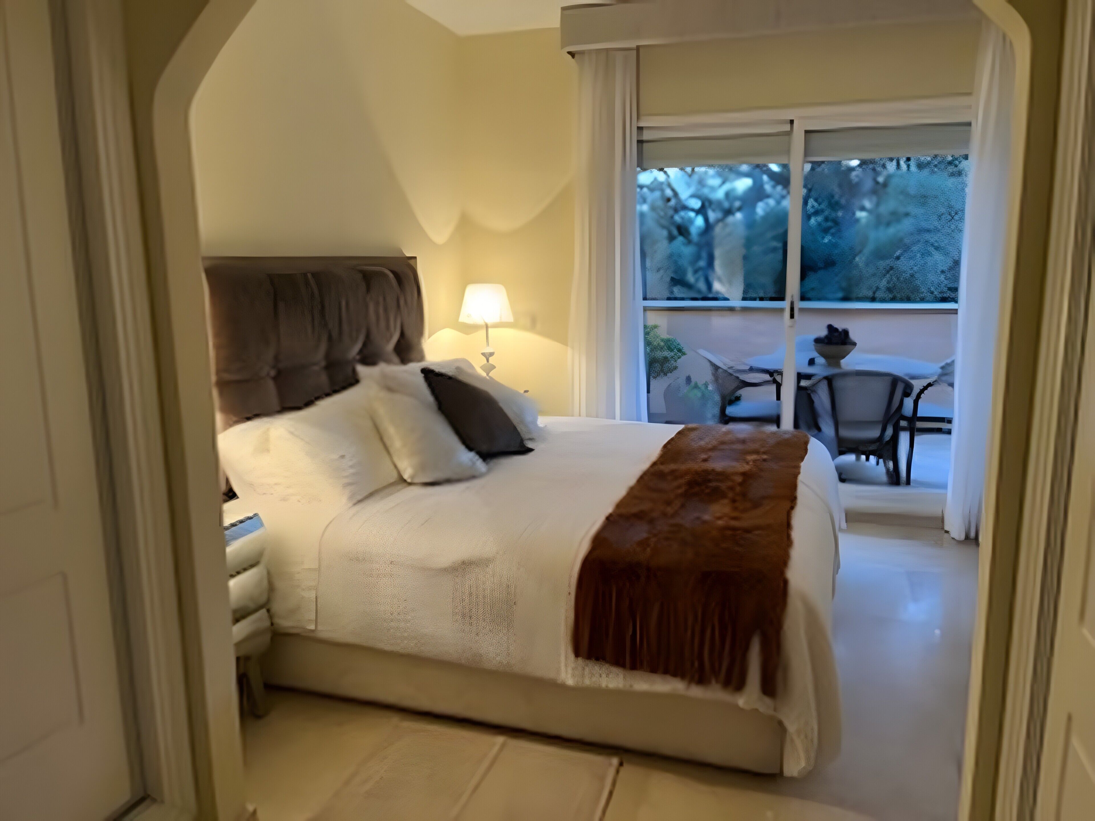 A bedroom with a bed, a nightstand, a lamp, and a view of an outdoor patio with chairs and a table.