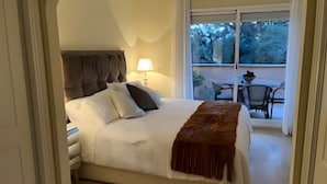 Une chambre à coucher comprenant un lit, une table de chevet, une lampe et donnant sur une terrasse extérieure avec des chaises et une table.