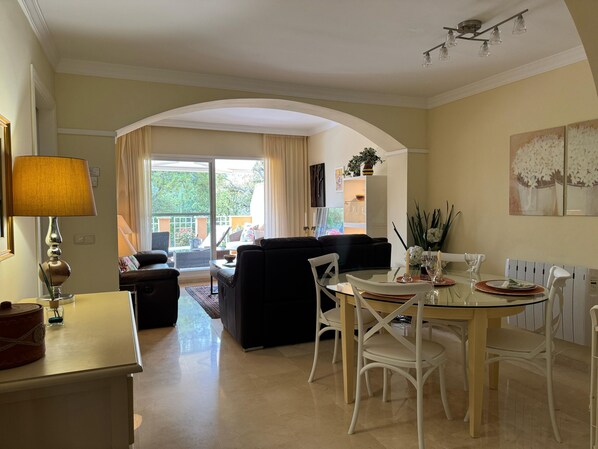 A living room with a sofa, a dining table with chairs, a lamp, and a view through a glass door to an outdoor area.
