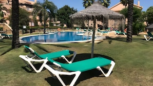 Un espace piscine avec des chaises longues, un parasol en chaume et des palmiers.