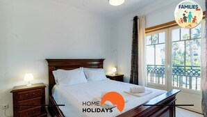 Un dormitorio con cama de madera, ropa de cama blanca, una mesita de noche con lámpara y ventana con vistas al verdor.