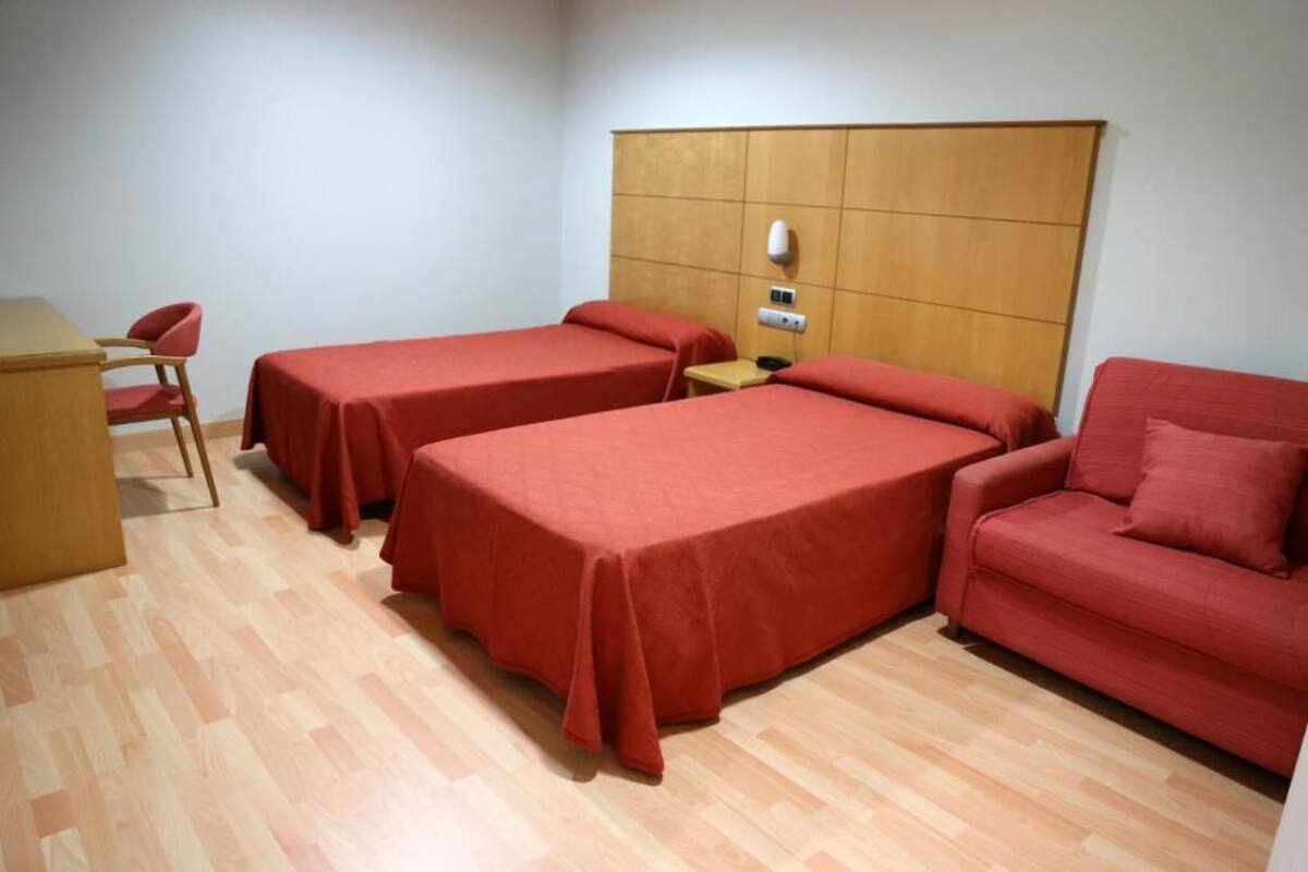 A room with two single beds, a wooden headboard, a small table, a red chair, and a red sofa.