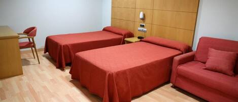 A room with two single beds, a wooden headboard, a small table, a red chair, and a red sofa.