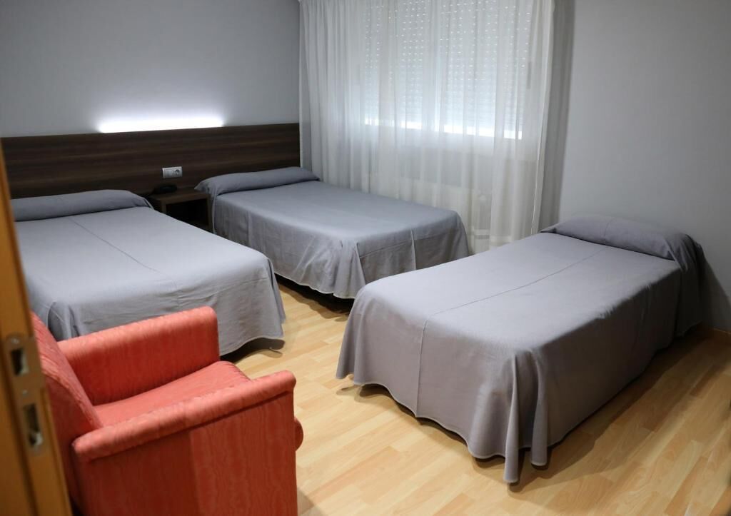 A hotel room with three single beds, a wooden floor, a red chair, and a window with sheer curtains.
