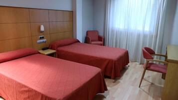 A hotel room with two single beds, a wooden headboard, a desk with a chair, and a window with curtains.