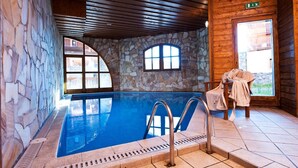Une piscine intérieure avec des murs en pierre et des poutres en bois au plafond, un banc en pierre et une vue sur l’extérieur.
