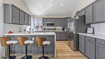 A modern kitchen with grey cabinets, a central island with bar stools, and a stainless steel refrigerator.
