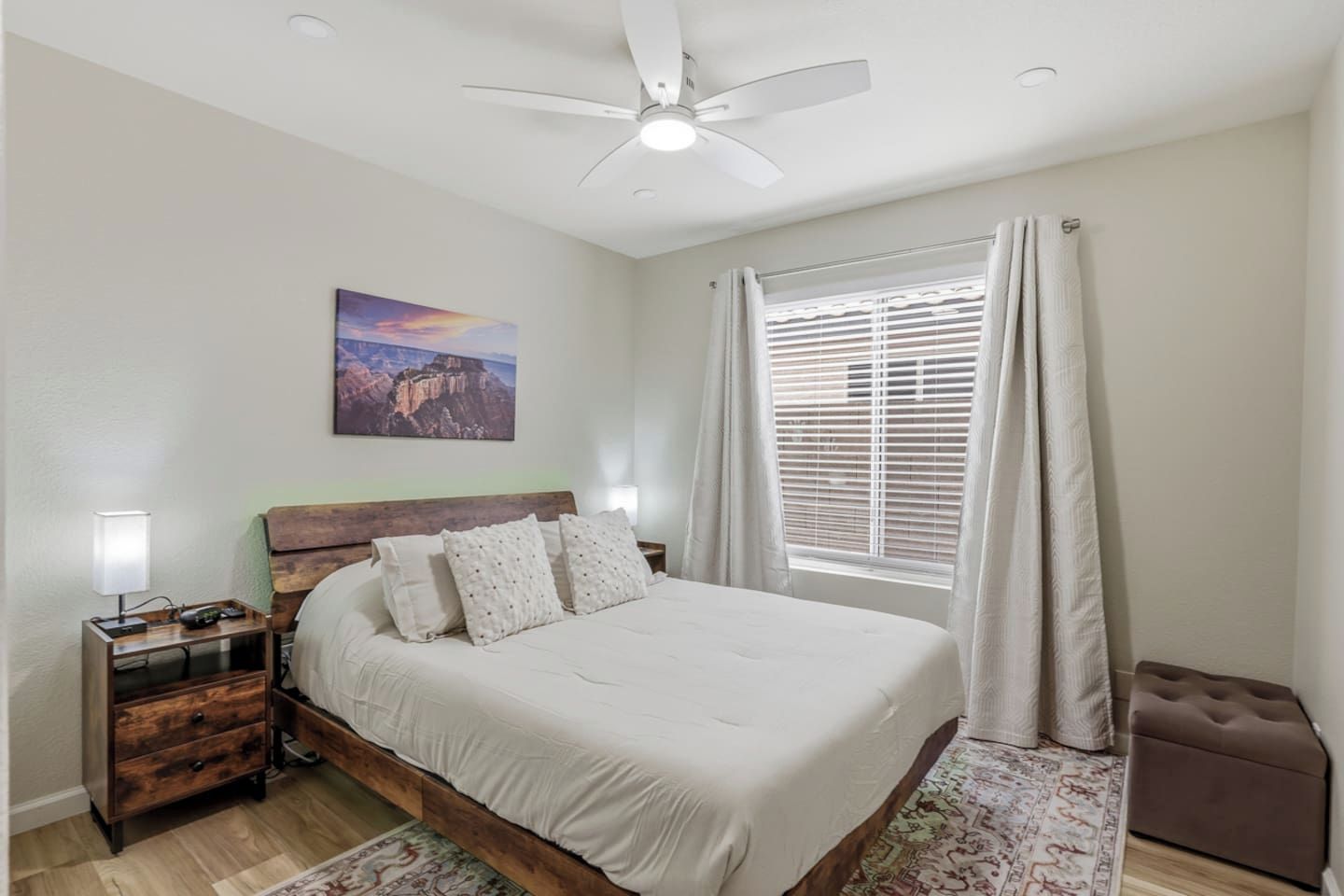 Une chambre à coucher comprenant un lit, une table de chevet, un bureau, un ventilateur de plafond, une fenêtre avec des rideaux et un tableau accroché au mur.
