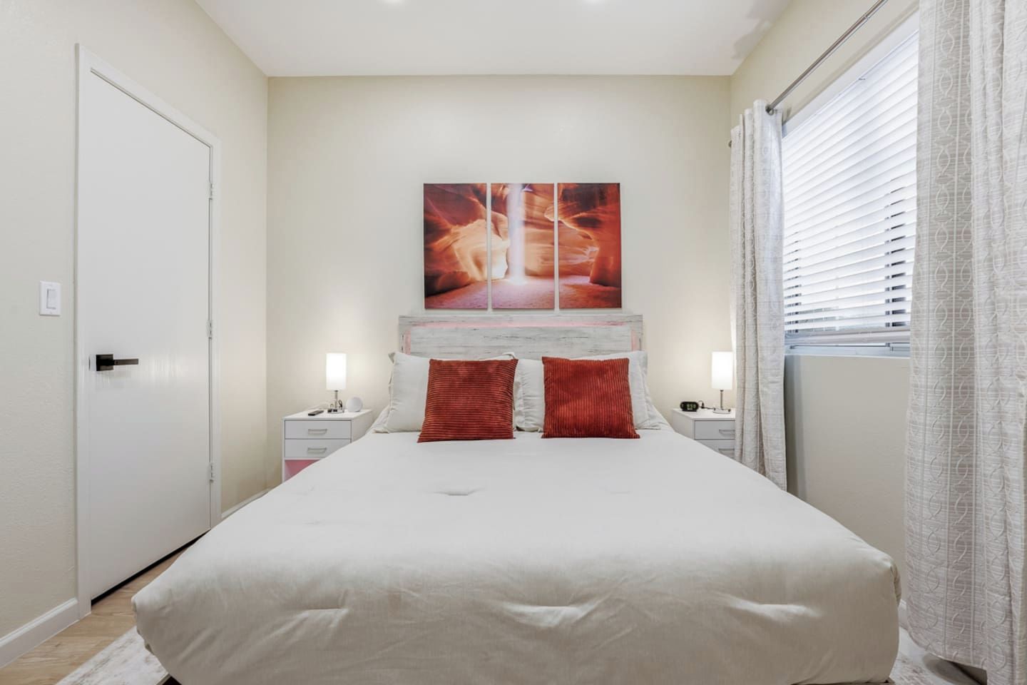 A bedroom with a bed, two nightstands, a large painting above the bed, and a window with blinds.
