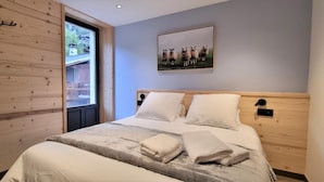 Ein Schlafzimmer mit hölzernem Kopfteil, einem Bett mit weißen Leinentüchern, einem Fenster mit Blick auf eine Holzterrasse und einem gerahmten Bild von Schafen an der Wand.