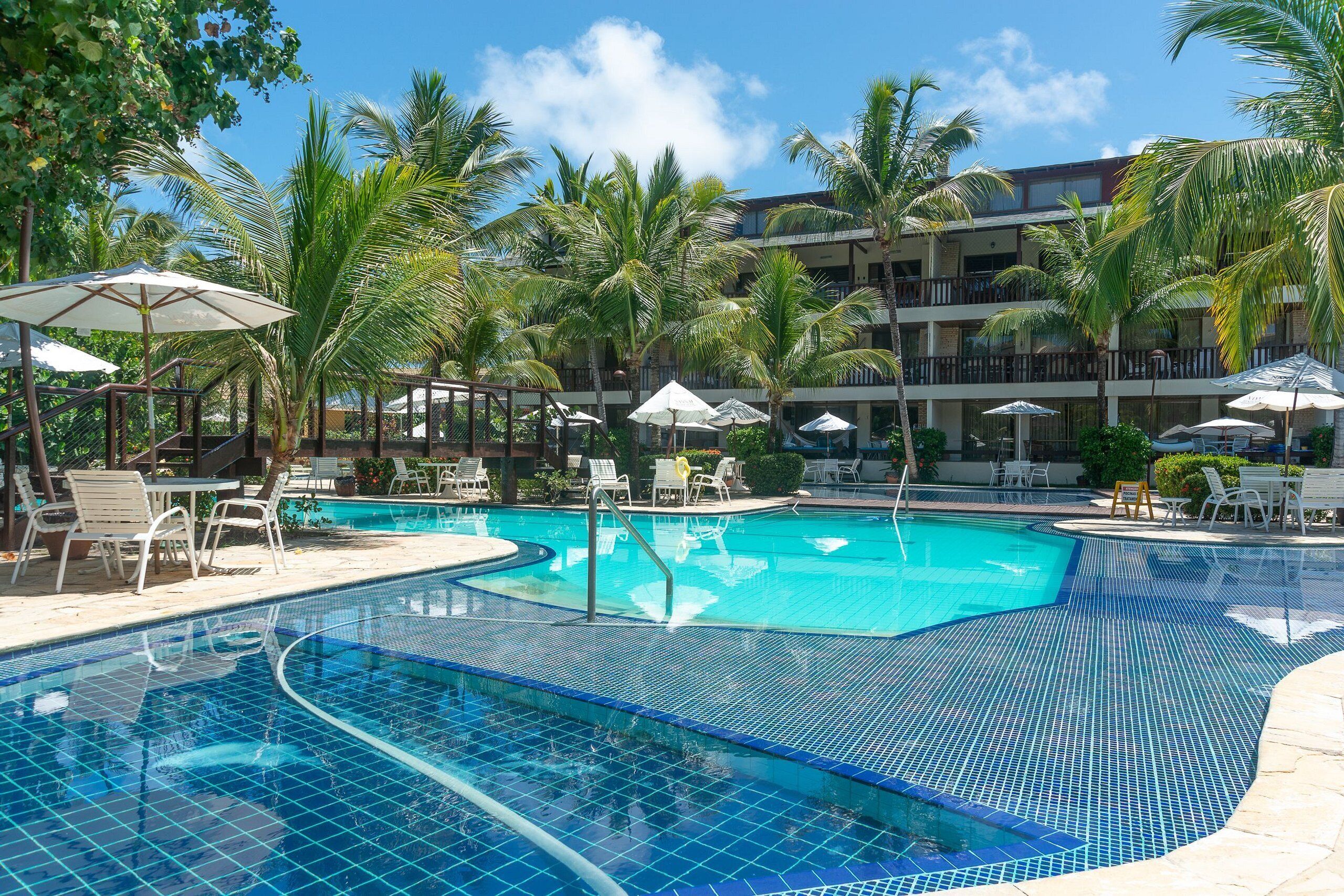 A swimming pool with tiled edges, surrounded by lounge chairs and umbrellas, with palm trees and a multi-story building in the background.