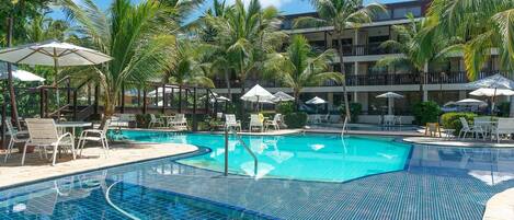 A swimming pool with tiled edges, surrounded by lounge chairs and umbrellas, with palm trees and a multi-story building in the background.