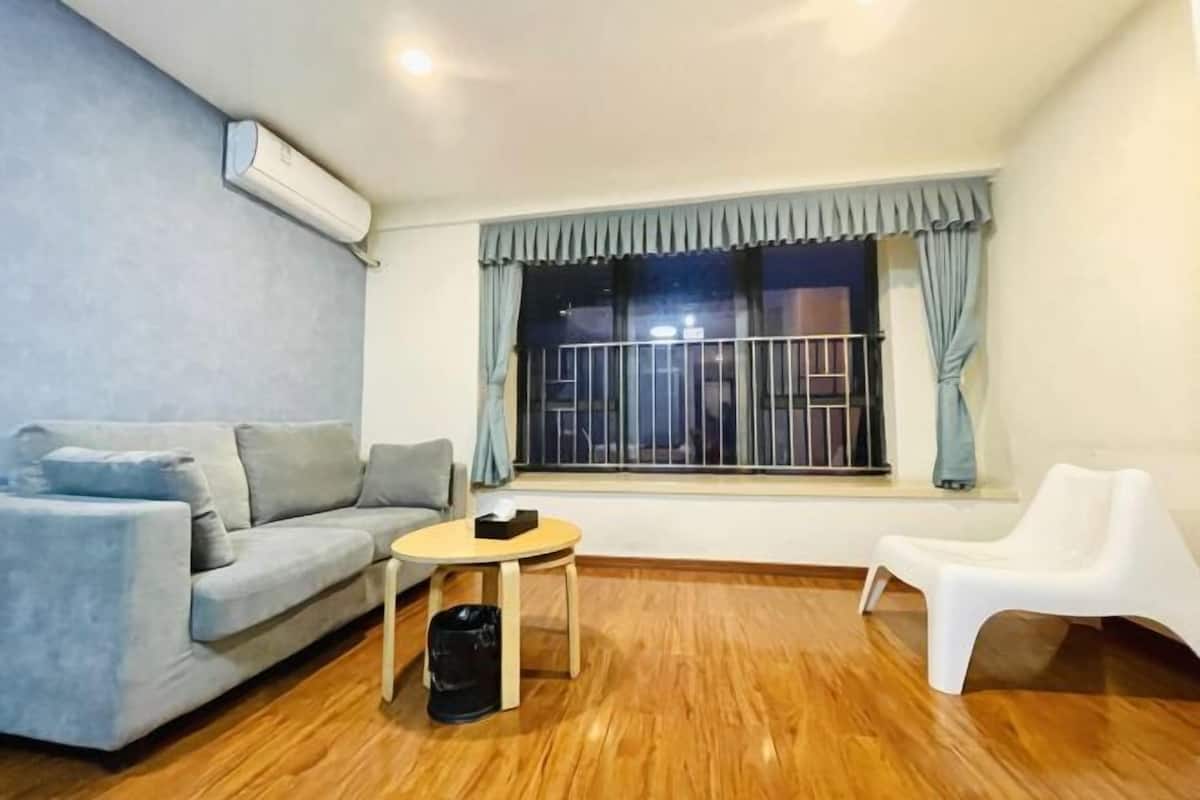 A room with a sofa, a small round table, a trash bin, a window with curtains, and a white chair.