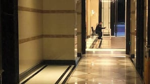 Een hotel lobby met een zwart-wit schakeerd motief op de vloer, een persoon zittend op een stoel en een glazen deur die naar een andere ruimte leidt.