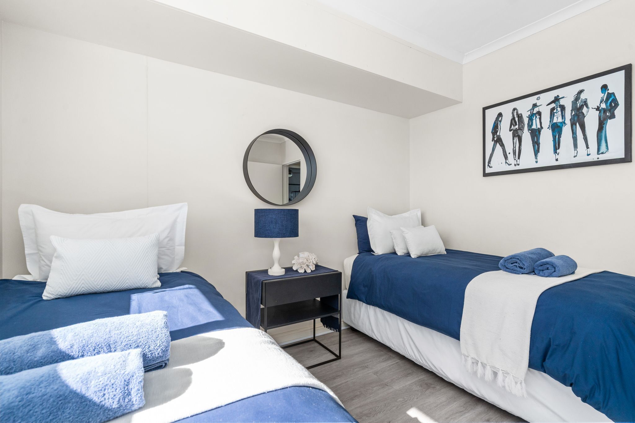 A hotel room with two beds, a nightstand with a lamp, a mirror, and a painting on the wall.
