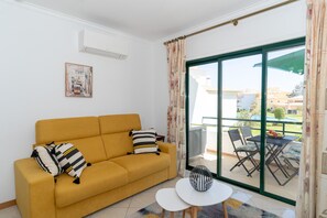 A yellow sofa with patterned pillows, a small white table, and a view of a balcony with outdoor furniture.
