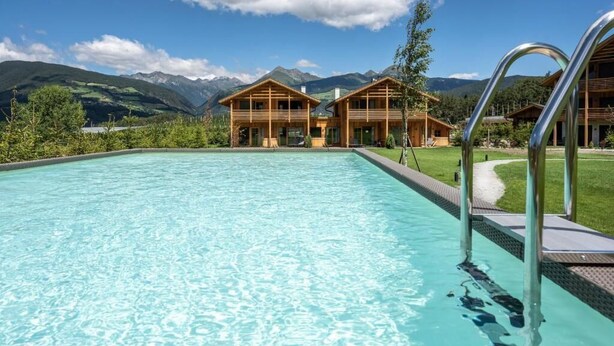 A swimming pool with wooden cabins in the background and mountains. - Kessler's Mountain Lodge (Naz Sciaves)