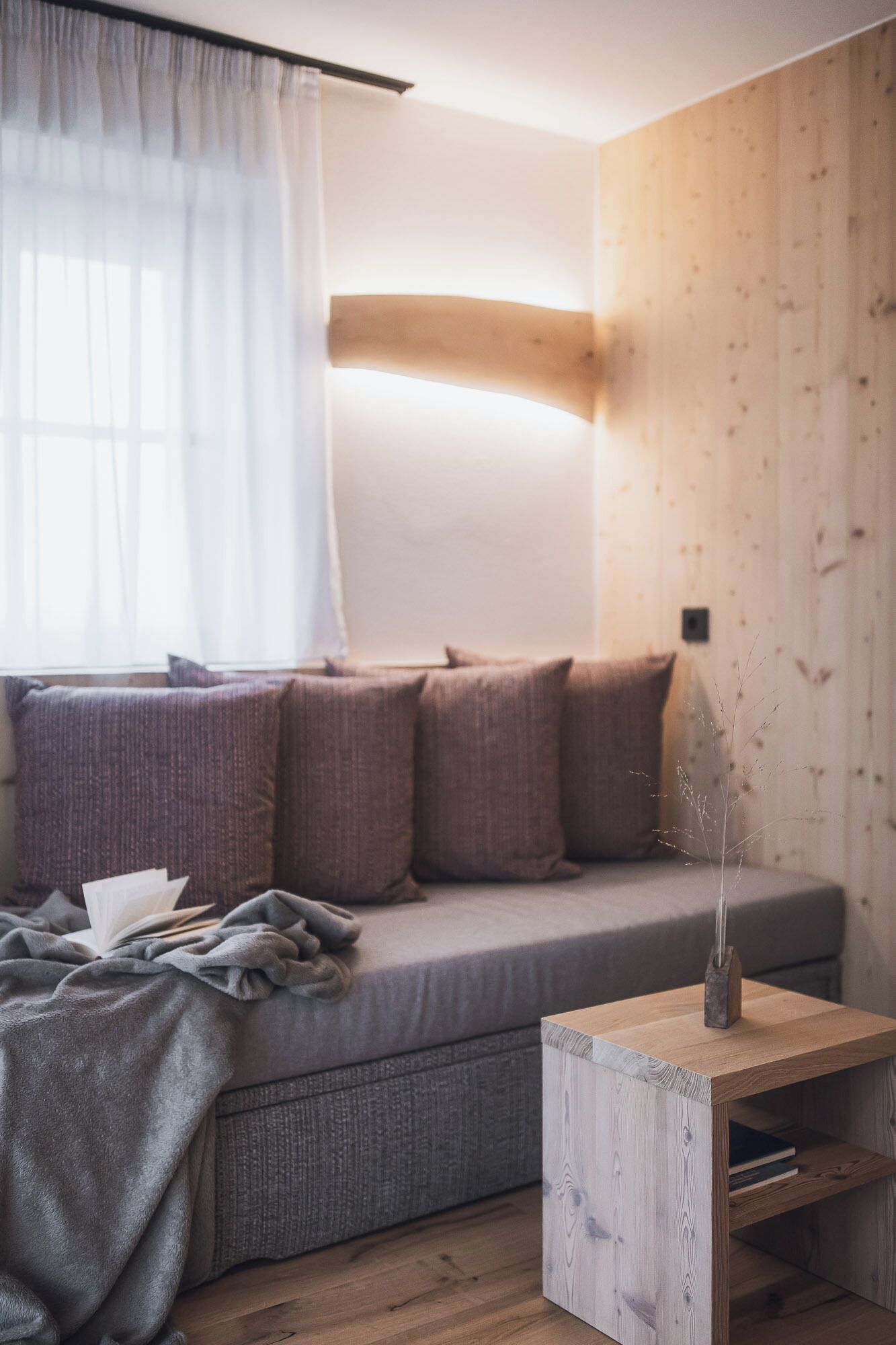 Una sala moderna con un sofá gris, una mesita de madera y una ventana con cortinas translúcidas.