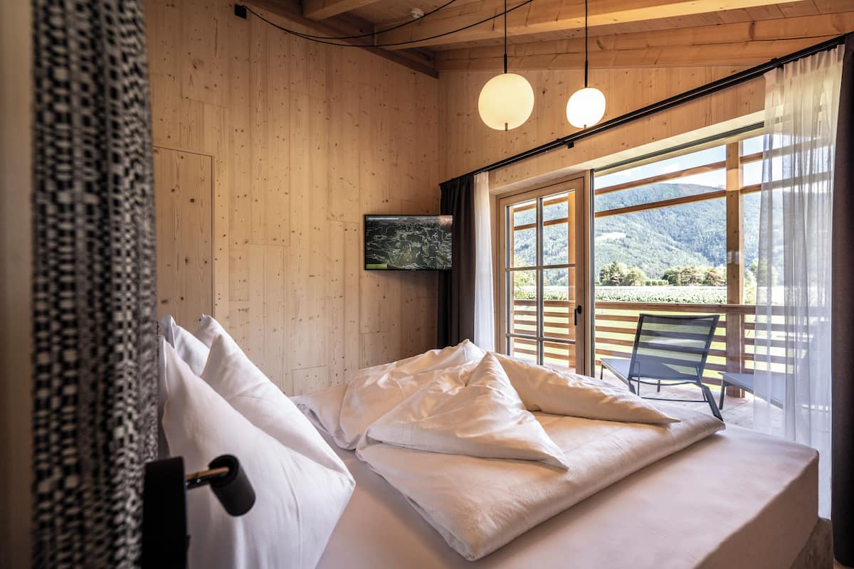 Ein Schlafzimmer mit Holzbalkendecke, einem Bett mit weißer Bettwäsche, einem an der Wand befestigten Fernseher und Blick auf einen Balkon mit einem Stuhl.