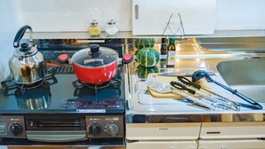 Una cocina con estufa, tetera, olla, tabla de cortar y diferentes utensilios.