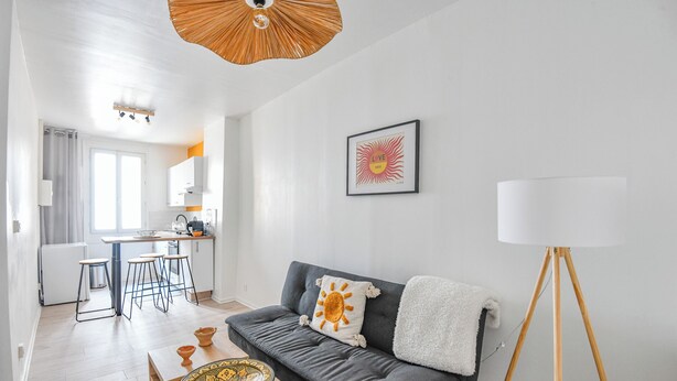 Una sala de estar moderna con un sofá gris, una mesa de centro de madera, una lámpara de pie y una cocina al fondo.