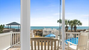 Une terrasse en bois avec des rampes blanches, un banc et une chaise, donnant sur une plage avec une piscine et des palmiers.