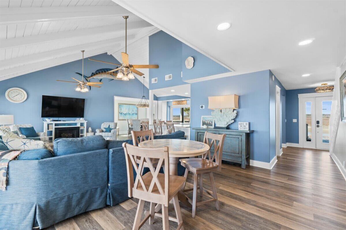 A spacious living room with a blue sofa, wooden dining table, and chairs. A ceiling fan and chandelier are visible.