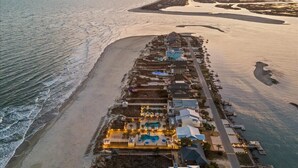 Vue aérienne d’un hôtel situé sur la côte, avec une longue plage, une piscine et plusieurs bâtiments dotés de balcons.