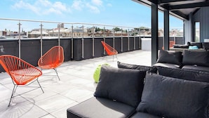 Une terrasse sur le toit avec un espace extérieur aménagé, comprenant un canapé et deux fauteuils, et offrant une vue sur le paysage urbain.