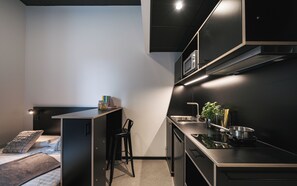 A compact kitchen with a countertop, sink, and stove. There is a small bar area with stools and a bookshelf. - Moliving - Design Hotels & Apartments - Free Gym (Neuss)