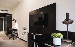Une chambre moderne dotée d’un téléviseur à écran plat fixé au mur, d’un bureau agrémenté d’une plante en pot, d’une chaise et d’une lampe.