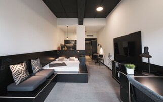 Una habitación de hotel moderna con sofá, cama, escritorio, televisor y kitchenette.