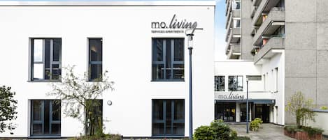 A modern serviced apartment building with a clean white facade, large windows, and a small outdoor garden area.