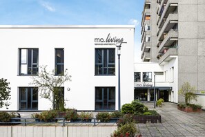 A modern serviced apartment building with a clean white facade, large windows, and a small outdoor garden area. - Moliving - Design Hotels & Apartments - Free Gym (Neuss)