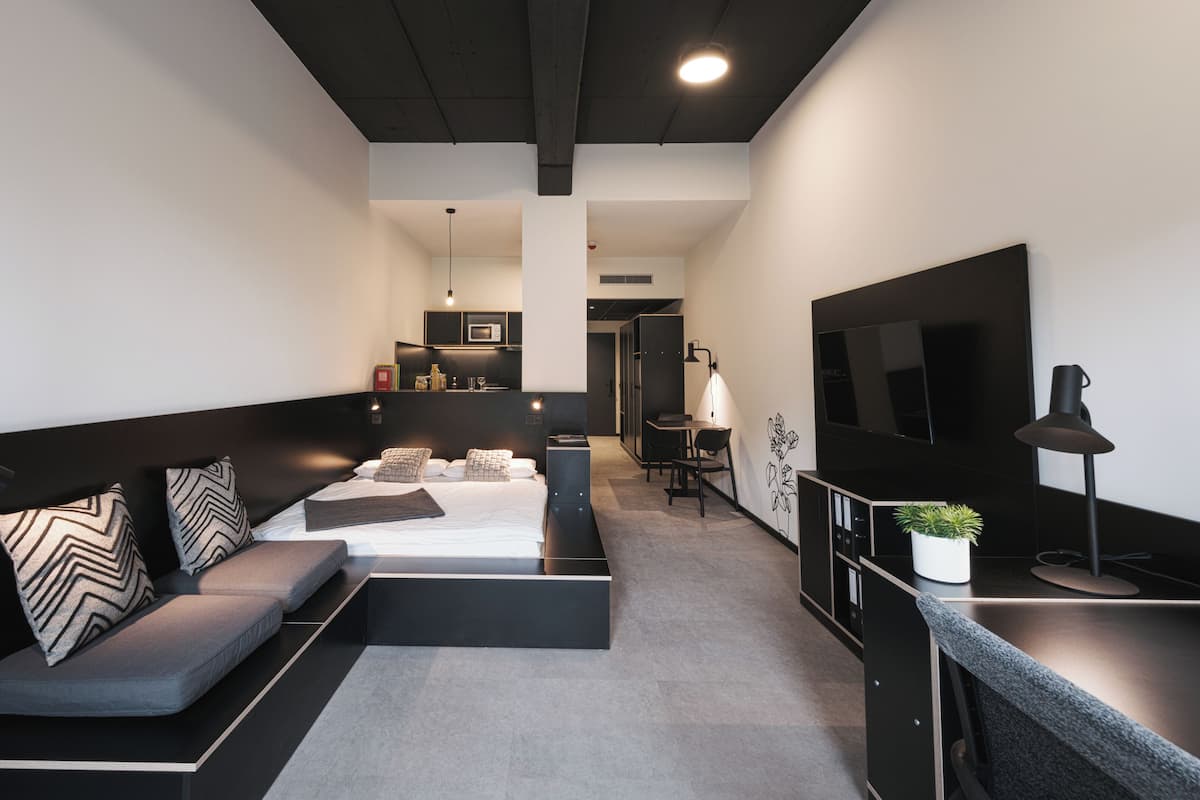 Una habitación de hotel moderna con sofá, cama, escritorio, televisor y kitchenette.
