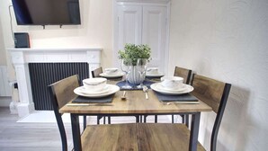 Uma sala de jantar com uma mesa de madeira posta para quatro pessoas, rodeada por cadeiras. Uma lareira é visível ao fundo e uma televisão de tela plana está montada na parede acima da mesa.