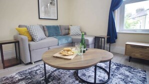 Uma sala de estar com um sofá, duas mesinhas laterais, uma mesa de centro e uma janela com cortinas.