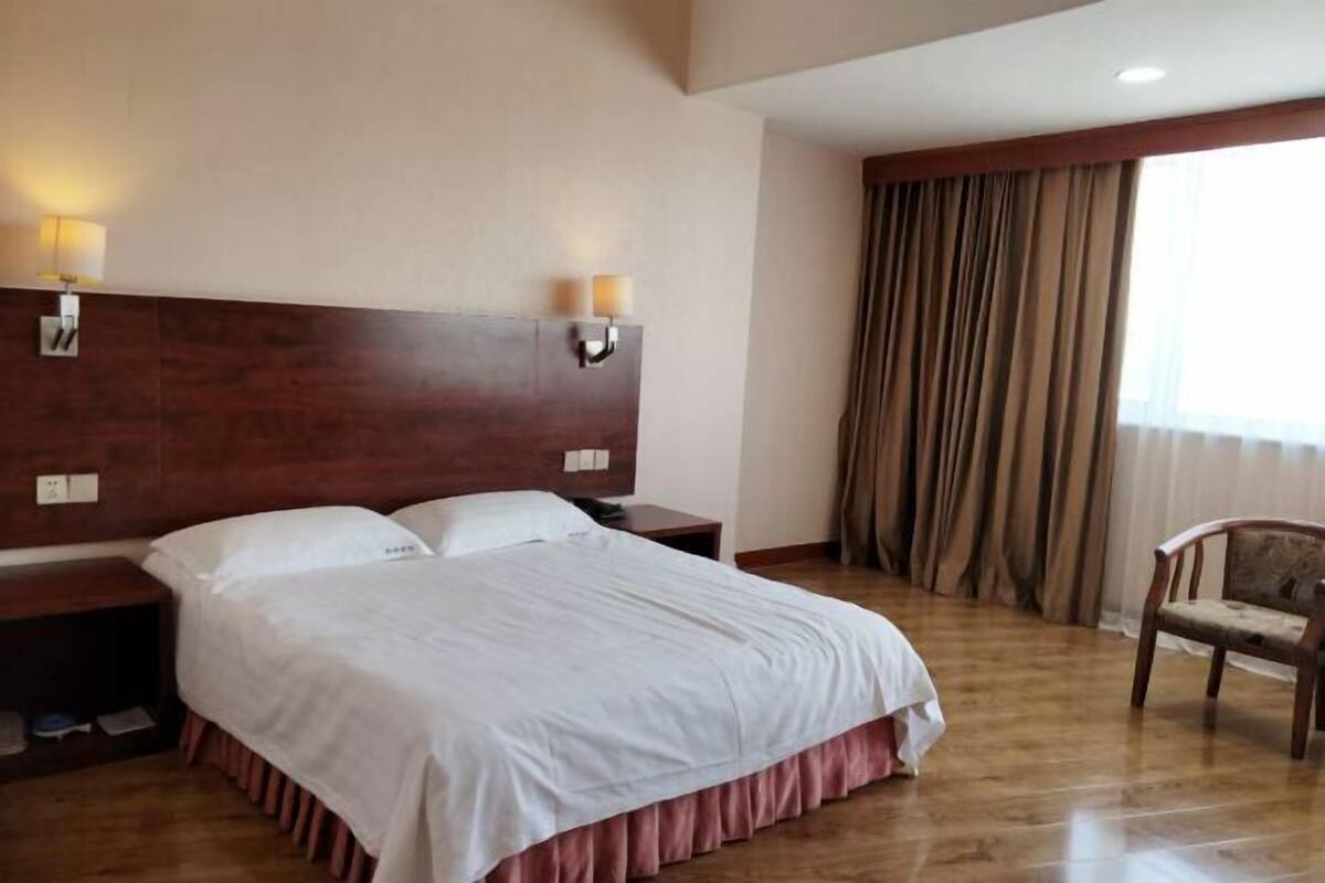 Une chambre d’hôtel avec une tête de lit en bois, un grand lit avec une literie blanche, deux lampes de chevet, une table de chevet en bois, une chaise et une grande fenêtre avec des rideaux.