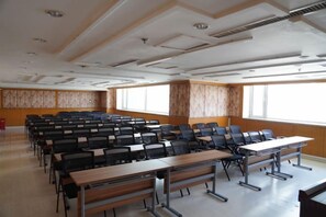 Een congreszaal met rijen stoelen en tafels, grote ramen en een wand van houten panelen.