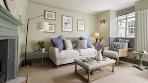 A living room with a sofa, armchairs, a coffee table, and a fireplace. There are framed pictures on the wall, a floor lamp, and a window with curtains. - Beautifully presented dog friendly - The Barley Apartment (Cirencester)