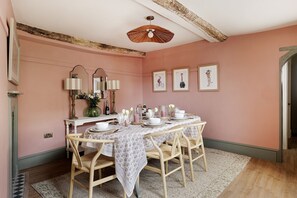 Une salle à manger avec une table ronde dressée pour quatre personnes, entourée de chaises en osier, d’une nappe blanche et d’œuvres d’art encadrées accrochées au mur.