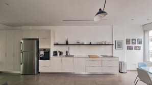 A modern kitchen with a stainless steel refrigerator, a built-in oven, and a sink. There are white cabinets, a black pendant light, and a window with a view of the outside.