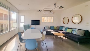 A modern living room with a dining table, chairs, a sofa, and a TV. There are large windows and a balcony visible.