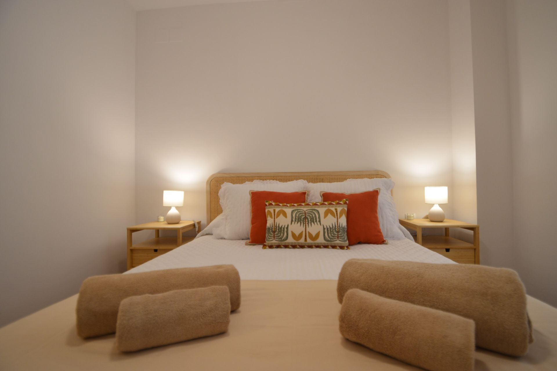 A neatly made bed with two pillows, two bedside tables with lamps, and two rolled towels on the bed.